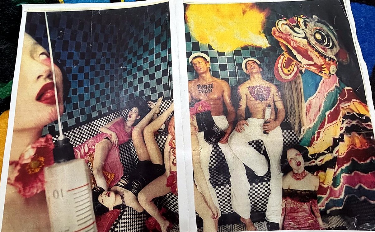 A magazine spread, torn out and creased: a giant syringe over red lips and two women on checkerboard tile, then a lion-dance head, a fire-breather and two tattooed men in sailor caps